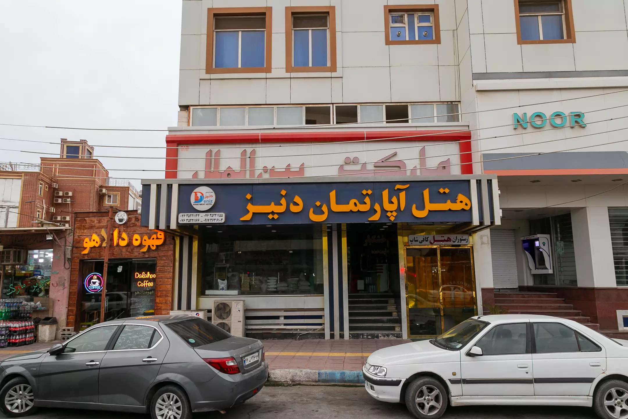 نمای ورودی هتل آپارتمان دنیز با تابلو و خودروهای خیابانی