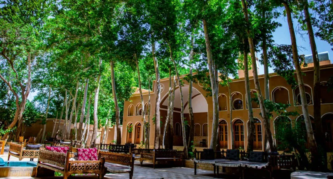 حیاط باغ خوشنویس با درختان بلند و تخت‌های سنتی