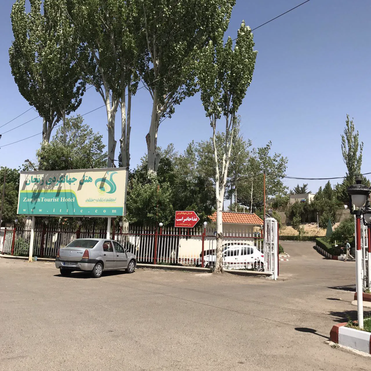 ورودی هتل جهانگردی با تابلو، درختان بلند و خودروها